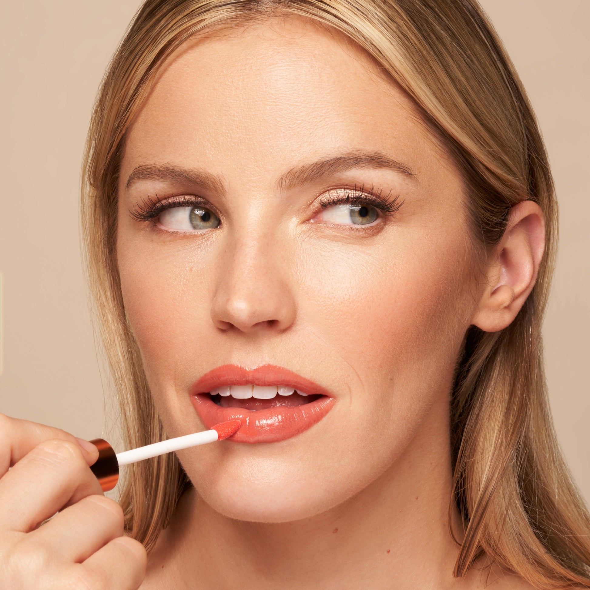 Glossy lip oil applied on lips in an orange-red shade, applicator near mouth