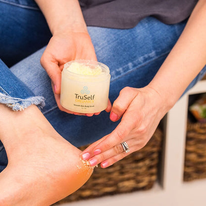 Hands scoop beige body scrub from a white-lidded jar; skincare texture on skin